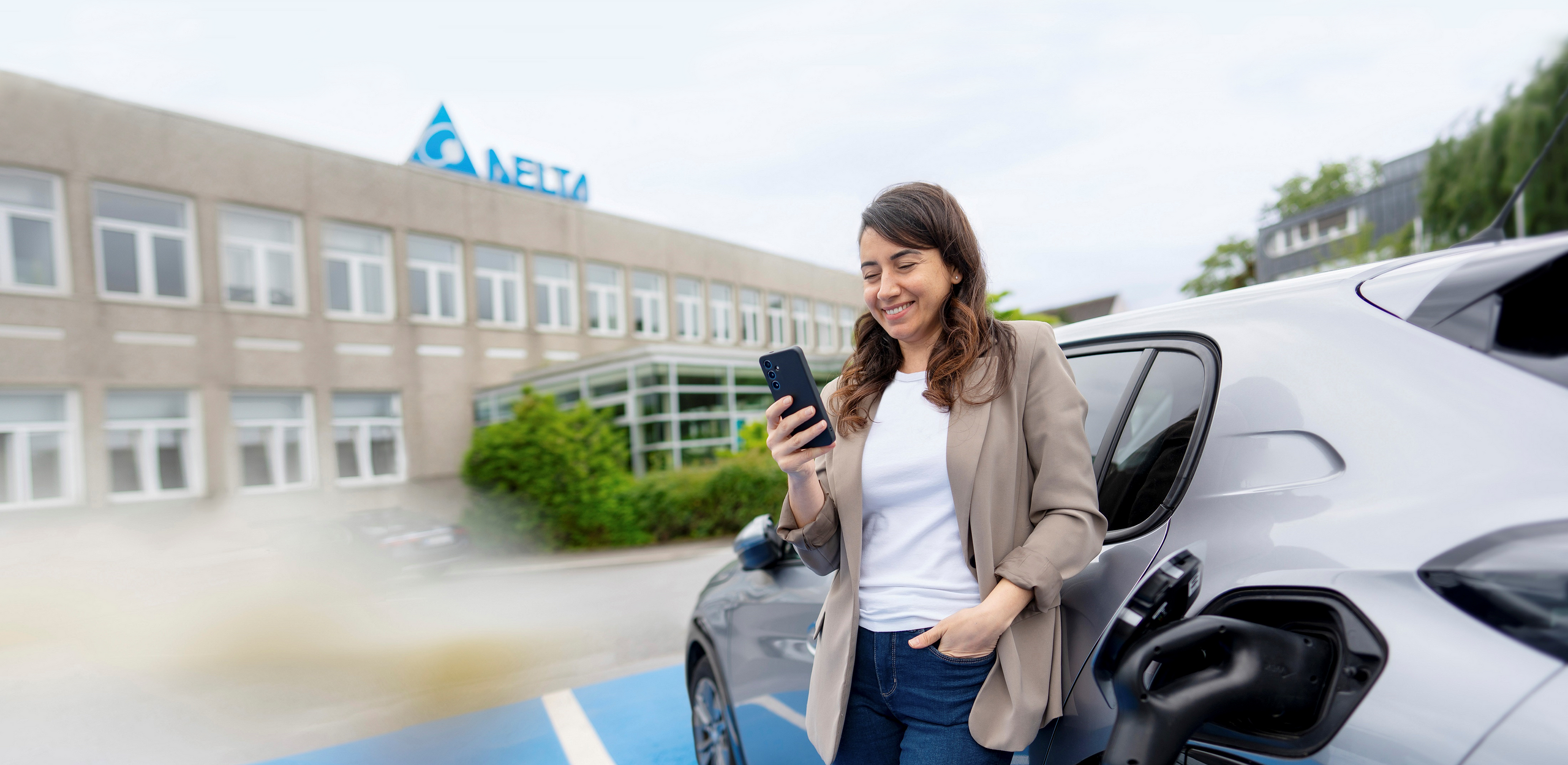 Eine Frau lädt ihr E-Auto auf und schaut dabei auf ein Smartphone, welches sie in der rechten Hand hält. Im Hintergrund ist ein Delta Bürogebäude zu sehen.