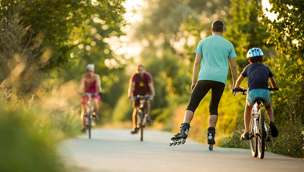 Personen und ein Kind fahren Fahrrad. Ein Mann fährt Inlineskater auf einem grünen Waldweg