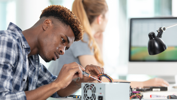 Ein dunkelhäutiger Student überprüft ein Bauteil und eine blonde Frau arbeitet am Computer.