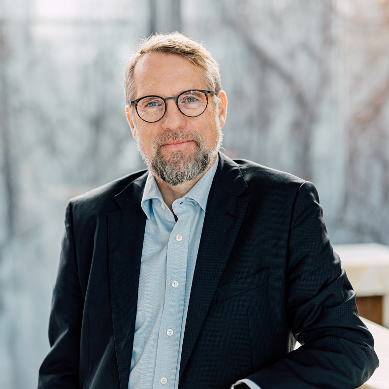 Andreas Kostrzewa A middle-aged man with glasses and a beard is wearing a black suit with a light blue shirt